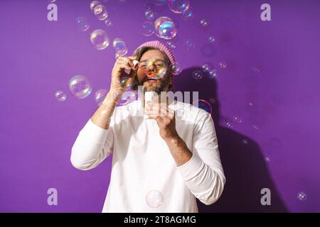 Uomo di mezza età spensierato che soffia bolle di sapone su sfondo viola Foto Stock