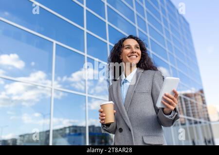 Una donna d'affari ispanica di successo va in ufficio, tiene in mano un caffè e un telefono, sorride e risolve i casi mentre si reca al lavoro, si trova per strada. Foto Stock