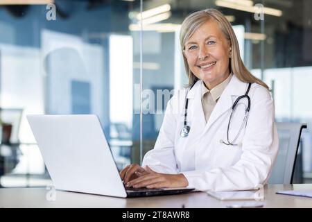 Ritratto di medico donna matura con esperienza, donna anziana dai capelli grigi in cappotto medico bianco sorridente guardando la macchina fotografica, consultando i pazienti a distanza, seduto al tavolo con il computer portatile all'interno della clinica. Foto Stock
