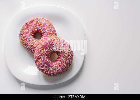 due ciambelle con glassa rosa e spruzzi colorati su un piatto bianco giacciono su un tavolo bianco, sulla destra c'è un posto per un'iscrizione. Alta qualità Foto Stock