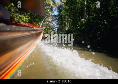 Acqua ondulata e gocce di spruzzi dietro la tradizionale barca a coda lunga in legno. Motoscafo sul canale fluviale tra le palme della Thailandia. Temi viaggi e. Foto Stock