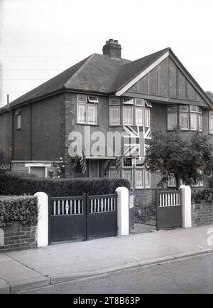 1953, storica, vista frontale di una casa suburbana semi-detatata costruita negli anni '1930, con una grande bandiera union Jack all'esterno, sotto la finestra, con bandiere più piccole sul portico, Inghilterra, Regno Unito, per celebrare l'incoronazione della Regina. Foto Stock