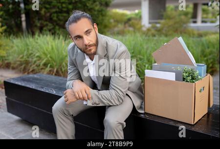 Sconvolge il giovane impiegato dell'ufficio seduto con una scatola di oggetti personali all'aperto Foto Stock