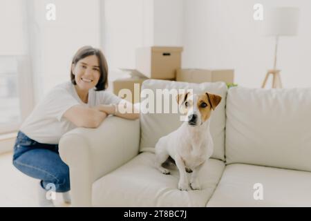 Donna sorridente e il suo cane su un divano bianco con scatole mobili in un soggiorno pieno di luce Foto Stock