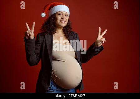 Bella donna incinta in elegante blazer grigio e cappello da Babbo Natale, sorride guardando la fotocamera, mostra il segno di pace con le mani, esprimendo un felice deposito Foto Stock