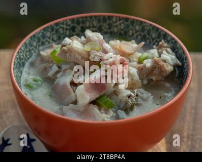 Riso bollito o porridge di riso con zuppa di costolette di maiale, prosciutto e sedano fresco in un recipiente di ceramica all'arancia. Cibo salutare per la colazione, vista dall'alto, spazio f Foto Stock