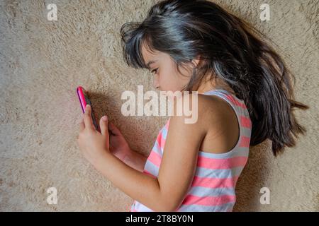 Bambina carina che usa uno smartphone. Ragazzo che gioca con il cellulare, sdraiato su un letto. Tempo libero. Tecnologia e concetto Internet. Bambino steso sul letto A. Foto Stock