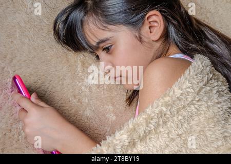 Bambina carina che usa uno smartphone. Ragazzo che gioca con il cellulare, sdraiato su un letto. Tempo libero. Tecnologia e concetto Internet. Bambino steso sul letto A. Foto Stock