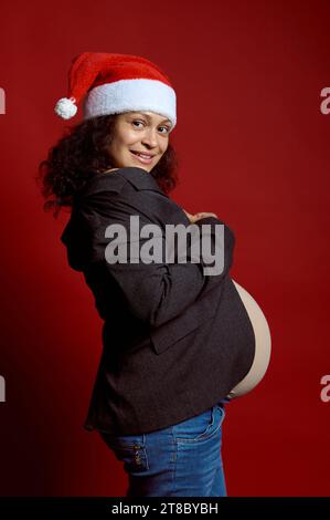 Ritratto autentico di una bella donna dai capelli ricci, una madre incinta a Babbo Natale, sorride guardando la macchina fotografica, esprime emozioni positive, isolato Foto Stock
