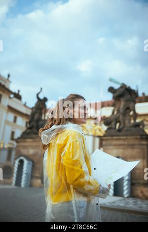 Felice donna turistica moderna in camicetta gialla e impermeabile a Praga Repubblica Ceca con mappa vicino al Castello di Praga. Foto Stock