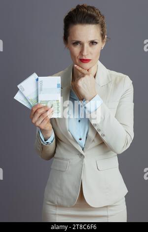 penosa lavoratrice alla moda di 40 anni in un abito da lavoro leggero con pacchetti di soldi da euro isolati sul grigio. Foto Stock