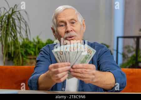 Pianificazione del budget familiare. Un uomo anziano felice che conta denaro contante calcola le bollette nazionali a casa. Il vecchio nonno anziano soddisfatto della pensione retributiva e risparmia denaro per i regali di vacanza programmati Foto Stock