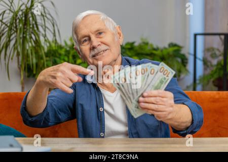 Pianificazione del budget familiare. Un uomo anziano felice che conta soldi in contanti puntando il dito sui dollari a casa. Il vecchio nonno anziano soddisfatto della pensione retributiva e risparmia denaro per i regali di vacanza programmati Foto Stock