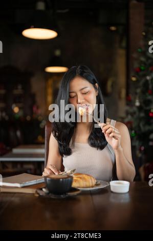 Elegante giovane donna asiatica che mangia croissant per colazione con latte caldo in un lussuoso salotto scuro. Colazione mattutina con caffè. Foto Stock