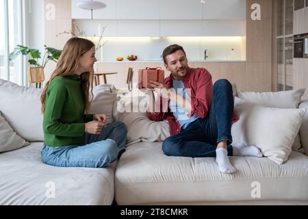 Donna gioiosa che regala una confezione regalo a sorpresa a un giovane che si congratula con il fatto di sedersi sul divano a casa. Foto Stock
