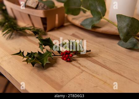 Su un tavolo di legno giace un ciuffo naturale di agrifoglio verde con bacche rosse. Simbolo di Natale. Pianta ornamentale per la realizzazione di bouquet invernali, ghirlande, interni d Foto Stock