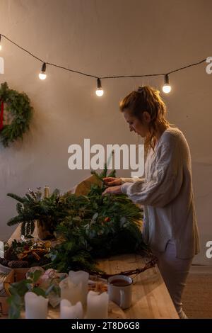 La giovane donna con maglione in maglia leggera sceglie aghi di pino verde naturale per la corona di Natale fatta a mano su un tavolo di legno. La ragazza millenaria fa la decoratio Foto Stock