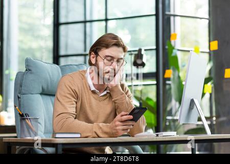 Un uomo stanco siede al tavolo con un telefono in mano, guarda tristemente lo schermo, sostiene la testa con la mano, brucia troppo lavoro, lavora in ufficio. Foto Stock