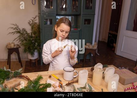 La bambina crea una corona di Natale fatta a mano a casa con materiali naturali. Autentico appartamento con interni scandinavi. Bambino che beve un tè caldo Foto Stock