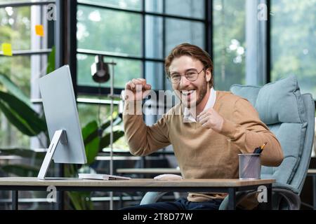 Un uomo d'affari molto felice alza le mani, si rallegra delle buone notizie, siede alla scrivania nell'ufficio di fronte al computer, guarda la fotocamera, sorride. Foto Stock