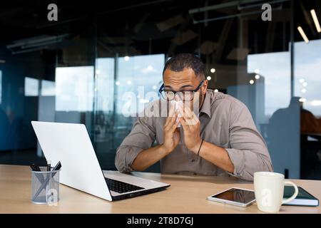 Uomo d'affari ispanico malato di una malattia virale in ufficio, ha un naso che cola, starnuti, allergie stagionali, siede a una scrivania, lavora malato. Foto Stock