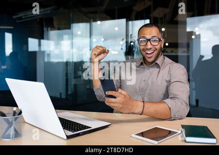 Gioioso uomo d'affari arabo che tiene un telefono in mano e solleva con gioia la mano, seduto a un tavolo in un atrio, felice di buone notizie, sorride e guarda la fotocamera. Foto Stock