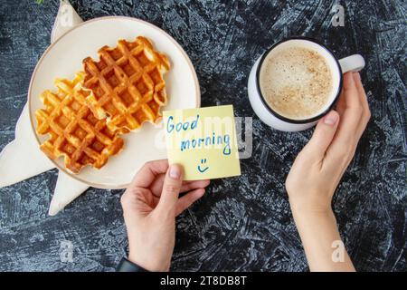 Vista dall'alto delle mani delle donne che tengono una nota con le parole GOOD MORNING accanto a una tazza di caffè e cialde viennesi fatte in casa macchiate su uno sfondo nero testurizzato. colazione a buffet Foto Stock