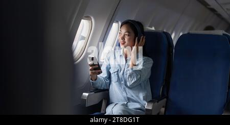 Giovane donna che ascolta la canzone preferita durante il volo in cabina di prima classe utilizzando lo smartphone, la donna si diverte a bordo dell'aereo e ascolta la musica in cuffia Foto Stock