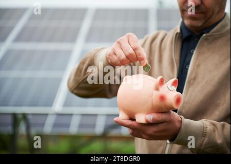 Investitore maschile che investe i suoi soldi in energie alternative. Vista ravvicinata dell'uomo che mette la moneta nel salvadanaio. Fare profitto salvando la natura. Foto Stock