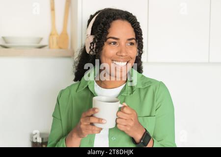 Donna nera che beve un caffè ascoltando musica nelle cuffie in cucina Foto Stock