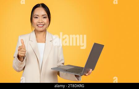 Felice e sicura giovane donna d'affari coreana in completo, con il computer, fare un gesto del pollice Foto Stock