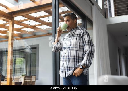 Un uomo birazziale anziano premuroso che beve caffè alla finestra di casa Foto Stock