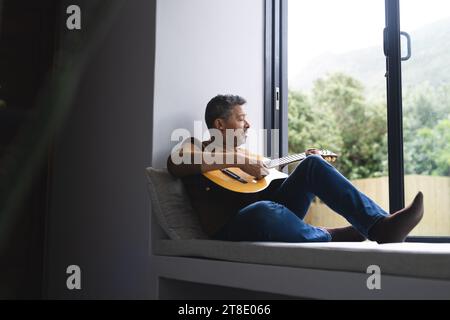 Felice uomo birazziale anziano che suona la chitarra alla finestra a casa Foto Stock