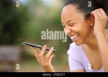 Felice donna nera che ride dettando un messaggio al telefono in un parco Foto Stock