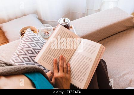 Foto orizzontale donna media adulta, caucasica, abbigliamento casual, seduta sul divano, accanto alla finestra, con un libro aperto in mano, con un tè. Foto Stock