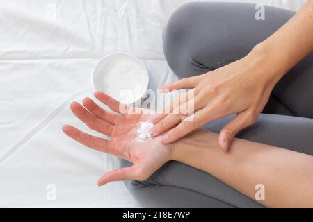 Foto orizzontale, mani di donna caucasica, vestita casualmente, con crema idratante sulle mani, seduta sul letto. Foto Stock