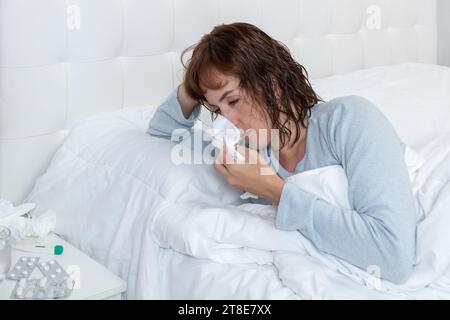 Foto orizzontale caucasica donna adulta, malata, fredda, che soffia il naso, a letto con pigiama blu a maniche lunghe. Foto Stock