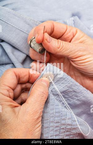 Foto ravvicinata verticale estrema, mani di donna caucasica adulta matura, mano che cuce un bottone con ago e ditale, su una camicia blu. Foto Stock