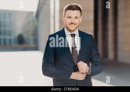 Uomo d'affari sicuro di sé che abbraccia la sua giacca in tuta all'aperto con una facciata moderna dietro l'hotel Foto Stock