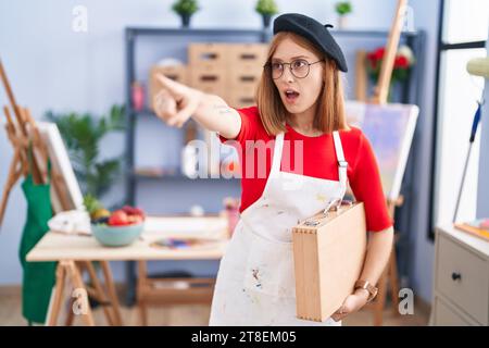Giovane donna rossa allo studio d'arte che porta un caso d'arte che punta con un dito sorpreso davanti, bocca aperta, espressione stupita, qualcosa sul davanti Foto Stock