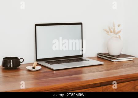 Computer portatile con schermo vuoto su un tavolo di legno all'interno della casa. Concetto di mockup sul luogo di lavoro. Lo sfondo dell'ufficio domestico con un computer portatile mock-up con spazio di copia Foto Stock