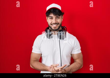 Uomo ispanico con barba che indossa cappello da giocatore e cuffie con le mani unite e le dita incrociate sorridendo rilassato e allegro. successo e ottimismo Foto Stock