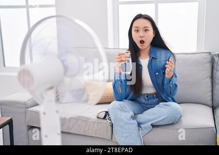 Giovane donna asiatica che beve un bicchiere d'acqua che si gode l'aria di un ventilatore spaventata e stupita con la bocca aperta per sorprendere e sfidare il volto incredulo Foto Stock