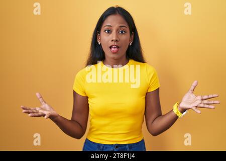 Giovane donna indiana in piedi su sfondo giallo sorridente allegra a braccia aperte come cordiale benvenuto, saluti positivi e fiduciosi Foto Stock