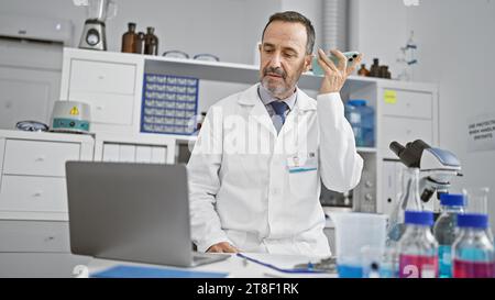 Uomo di mezza età dai capelli grigi ispanici, scienziato di laboratorio, impegnato nella ricerca scientifica, ascoltando un messaggio vocale sul suo smartphone in laboratorio Foto Stock