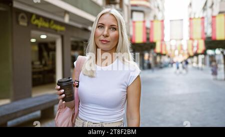 Una giovane donna bionda rilassata e bella che tiene una tazza di caffè da asporto in una strada soleggiata della città, esprimendo il suo stile di vita urbano all'aperto. Foto Stock