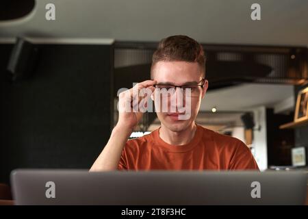 Un giovane sorridente che regola gli occhiali quando legge le e-mail sul laptop o cerca insetti Foto Stock