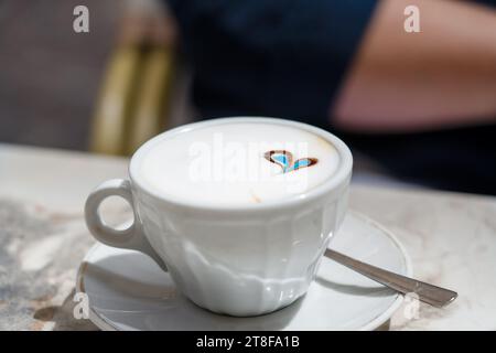 una tazza di caffè con parte superiore in schiuma di latte bianca e un disegno a forma di cuore blu Foto Stock