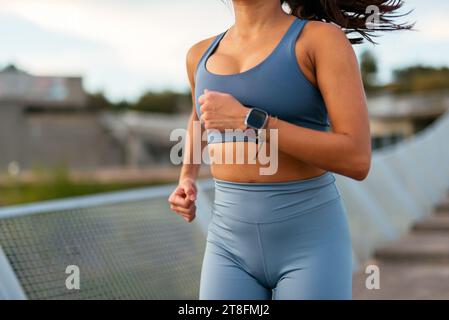 Una donna latinoamericana che indossa abbigliamento sportivo fa jogging con uno smartwatch, mostrando uno stile di vita sano in un ambiente urbano. Foto Stock