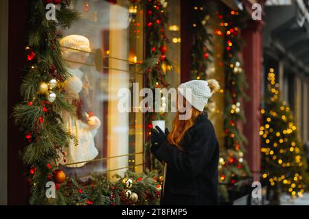 Una giovane donna con un cappello in maglia bianca e un cappotto nero tiene una tazza da caffè mentre si osserva una vetrina natalizia decorata con luci e decorazioni Foto Stock